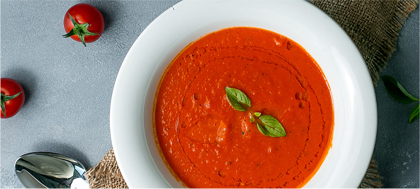 Tomato soup garnished with basil and olive oil, surrounded by tomatoes and textures.